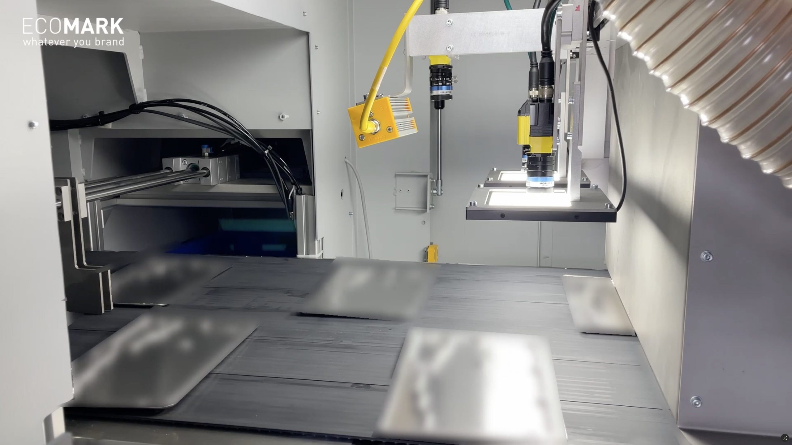 Nahaufnahme einer automatisierten Maschine mit einem Roboterarm, der Metallplatten auf einem Förderband markiert oder graviert, wobei Drähte und ein gelber Sensor sichtbar sind. Das ECOMARK-Logo erscheint in der oberen linken Ecke. eco-mark ecomark laserkennzeichnung laserbeschriftung industrie