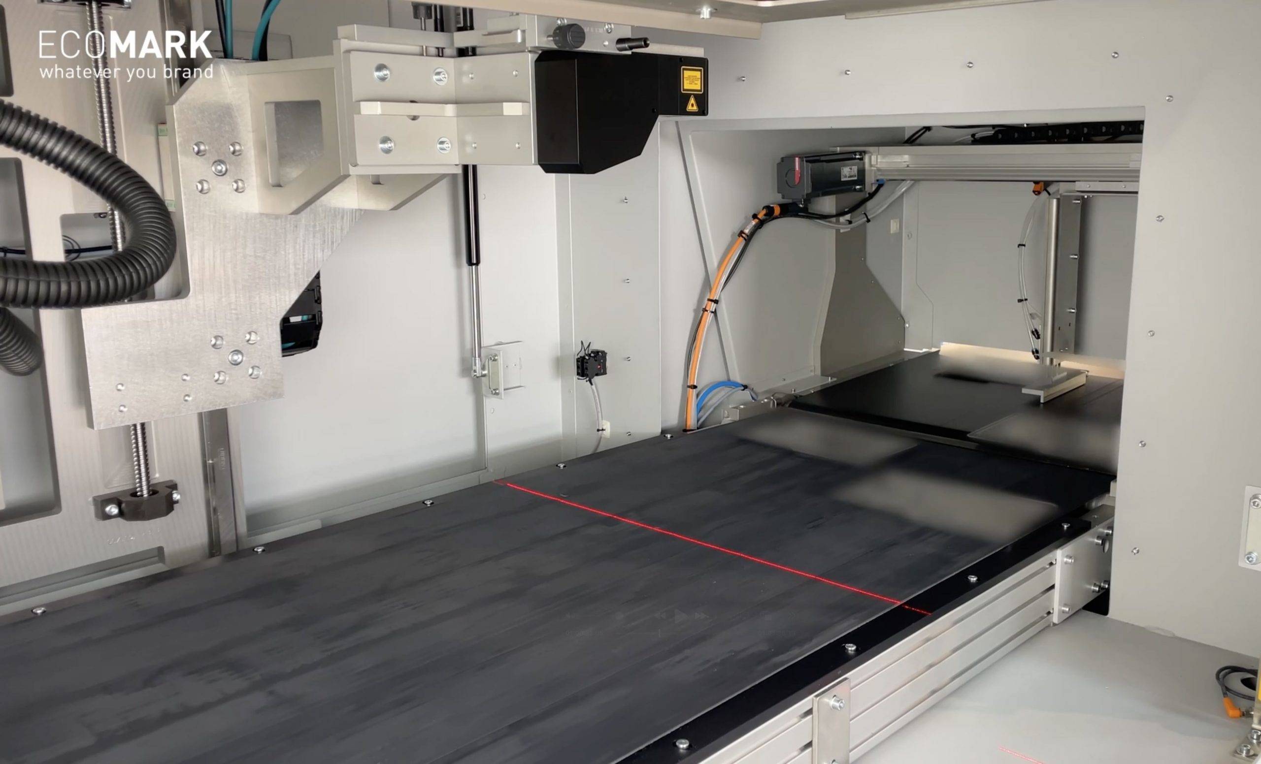 Nahaufnahme einer industriellen Laserbeschriftungsmaschine mit einem Förderbandsystem, auf dem rote Führungslinien und verschiedene Metall- und Elektronikkomponenten in einem sauberen, modernen Gehäuse zu sehen sind. eco-mark ecomark laserkennzeichnung laserbeschriftung industrie