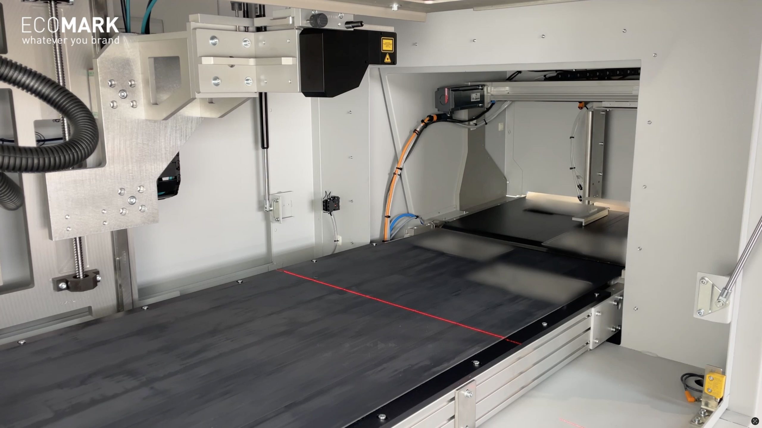 Nahaufnahme einer industriellen Lasermarkierungs- oder -graviermaschine mit einem Fördersystem in einer sauberen, modernen Anlage. Rote Richtungslaser werden auf die dunkle Förderbandoberfläche projiziert. eco-mark ecomark laserkennzeichnung laserbeschriftung industrie